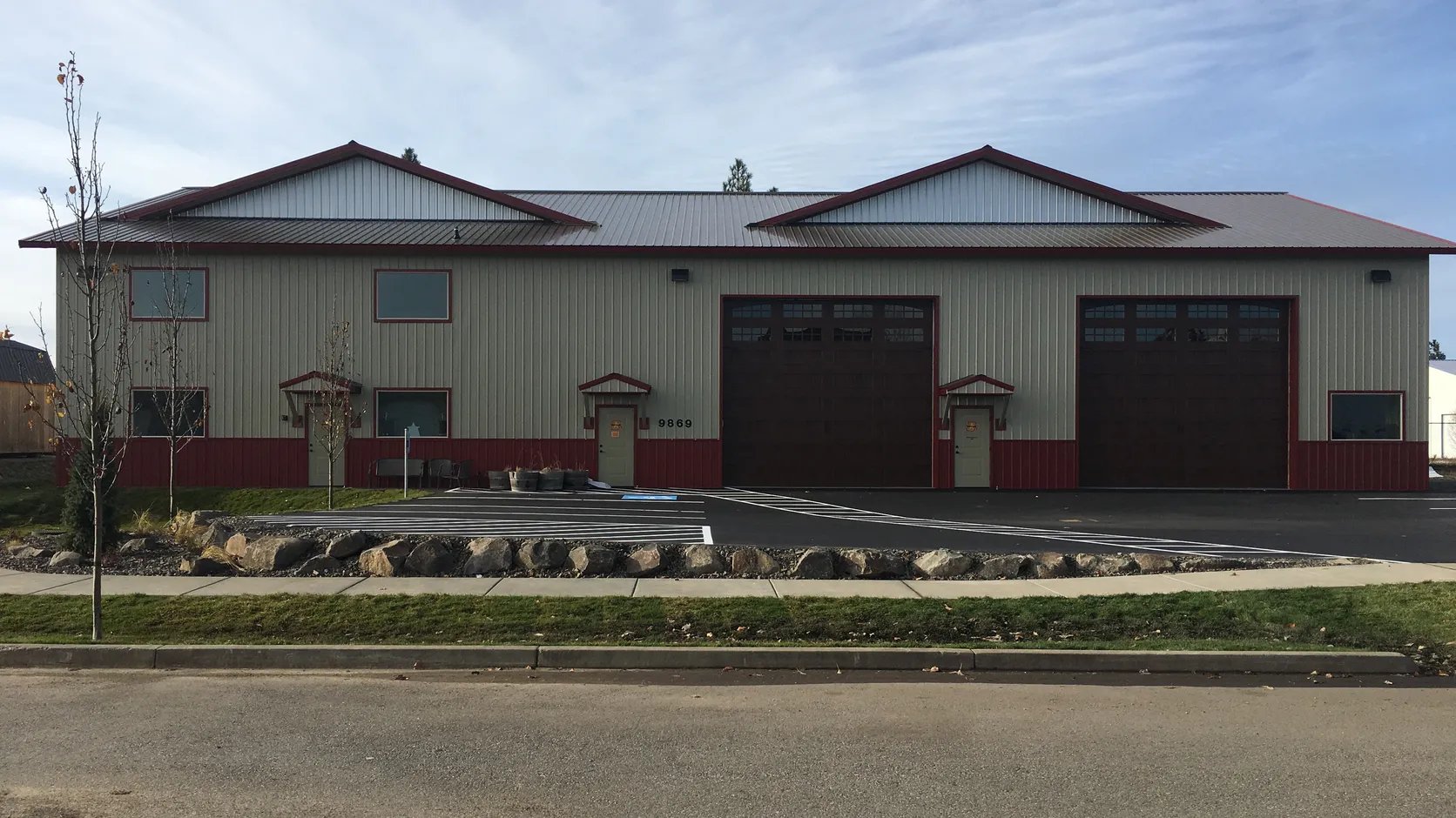 Completed commercial building with light gray metal siding, red wainscot and trim, two tall overhead bay doors, and a striped asphalt parking lot