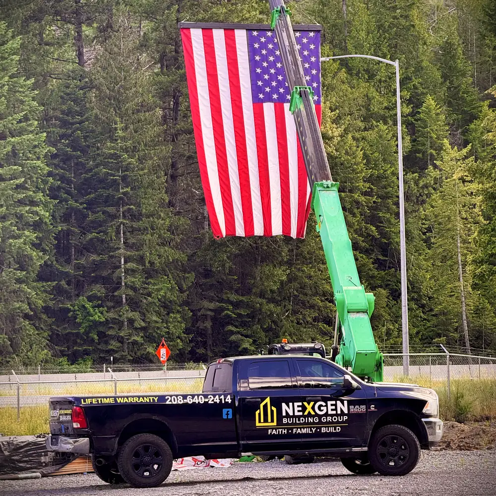 NEXGEN Building Group pickup truck on a job site, with a large American flag flown from a telehandler boom against the treeline